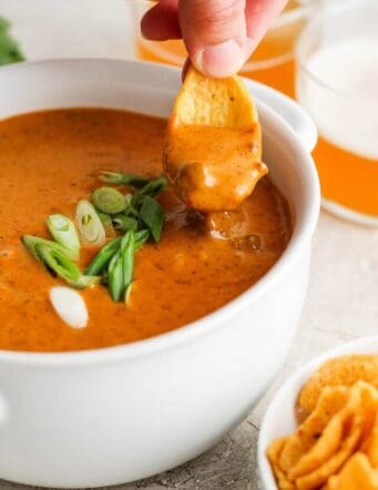 A hand uses a chip to scoop up chili cheese dip from a white bowl.