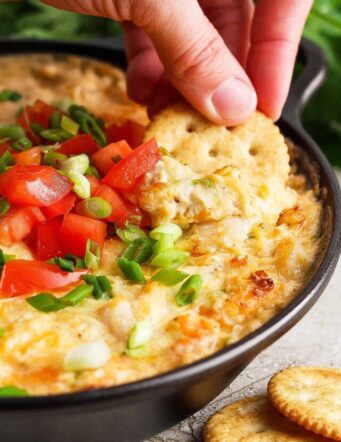 A hand dips a cracker into a tomato-topped bowl of shrimp and crab dip.