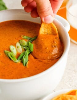 A hand dips a chip into a bowl of chili cheese dip.