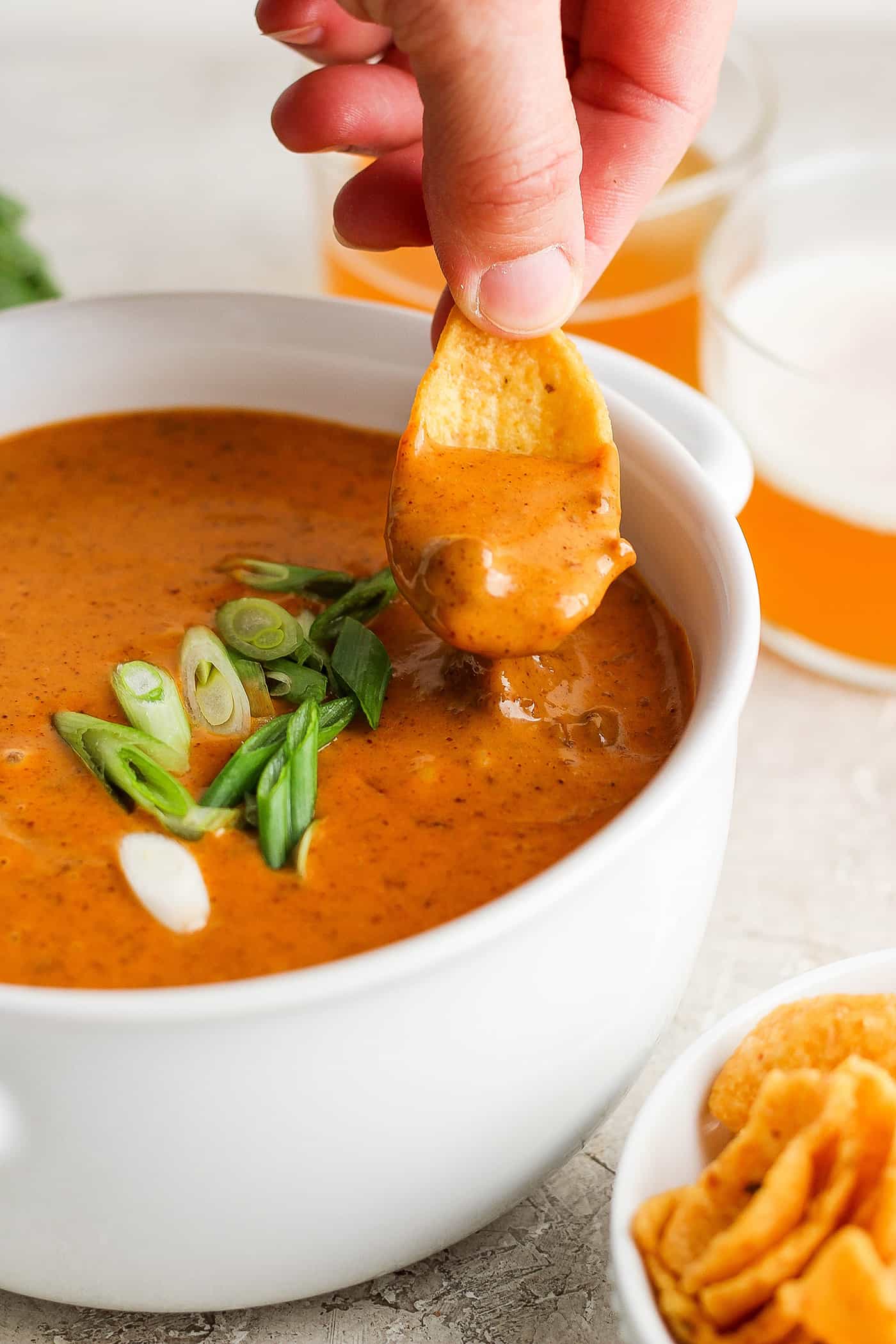 A hand uses a chip to scoop up chili cheese dip from a white bowl.