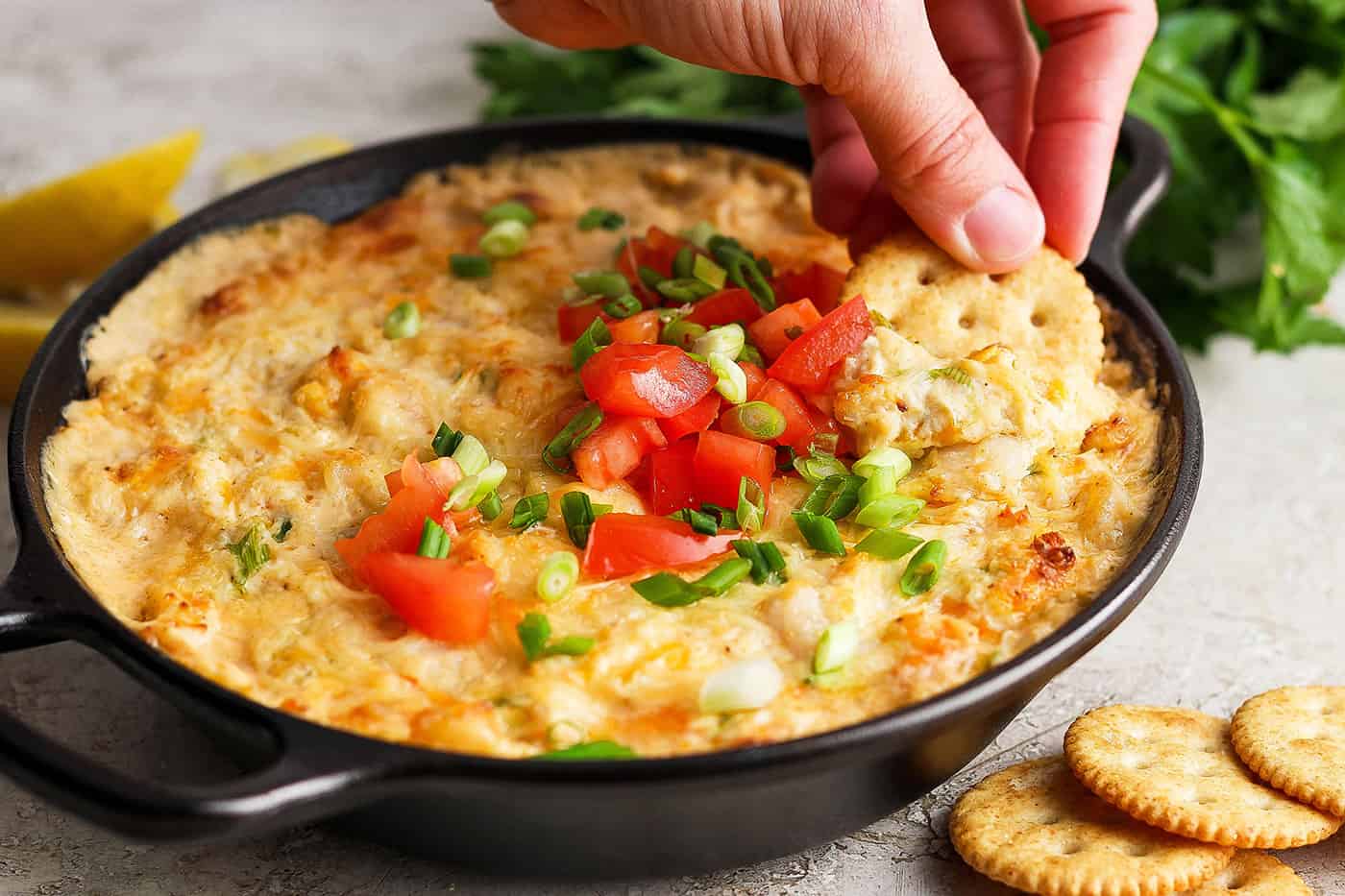 A hand dips a cracker into a tomato-topped bowl of shrimp and crab dip.