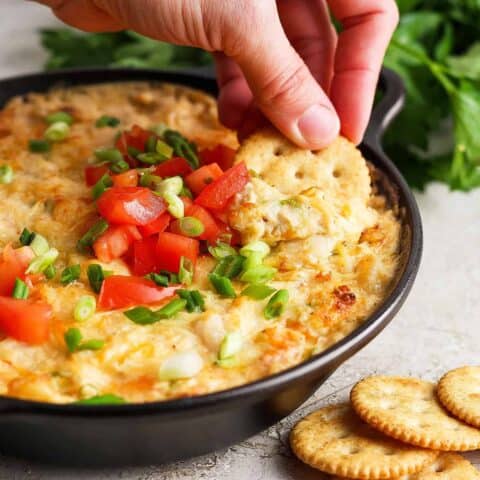 A hand dips a cracker into a tomato-topped bowl of shrimp and crab dip.