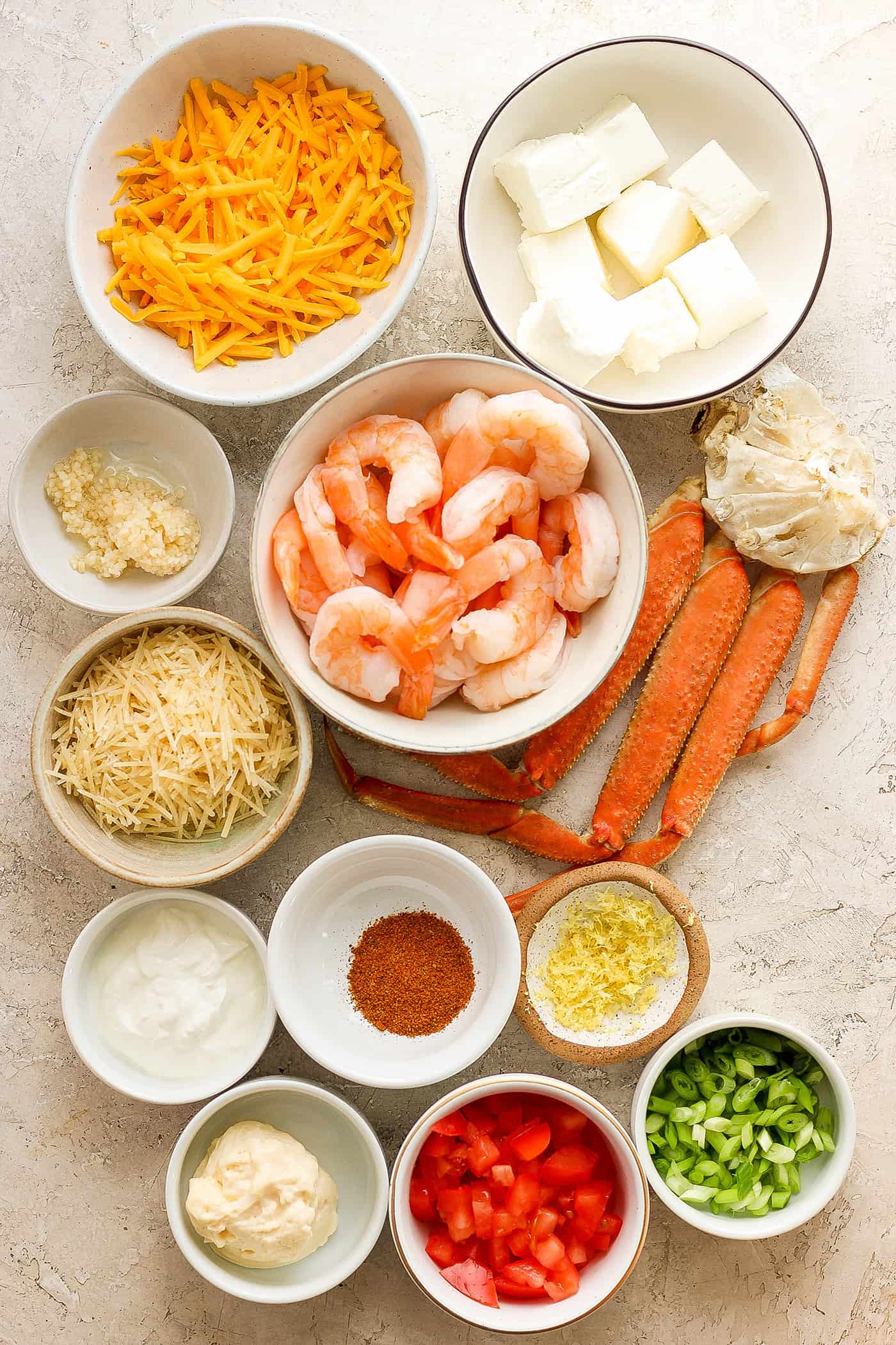 The ingredients for shrimp and crab dip are shown on a white background: shrimp, crab, garlic, shredded cheddar cheese, shredded parmesan cheese, sour cream, cream cheese, garlic, tomatoes, green onions.