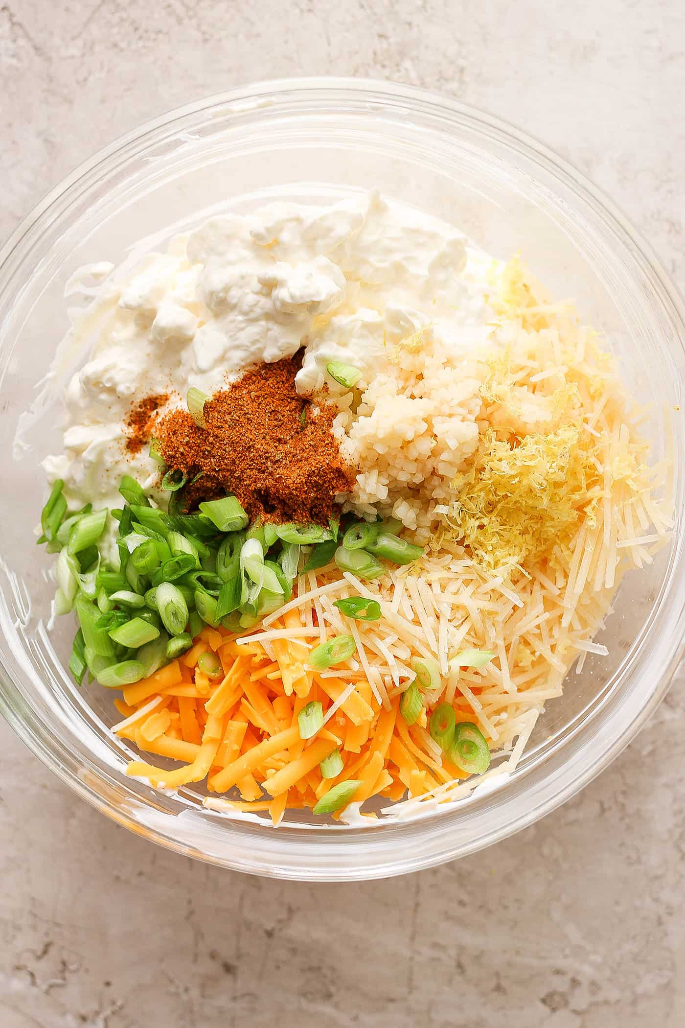 Ingredients for shrimp and crab dip including shredded cheddar, old bay, green onions, cream cheese and sour cream are shown in a bowl.