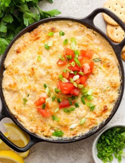 A cast iron pan holds baked shrimp and crab dipped garnished with chopped tomatoes and green onions.