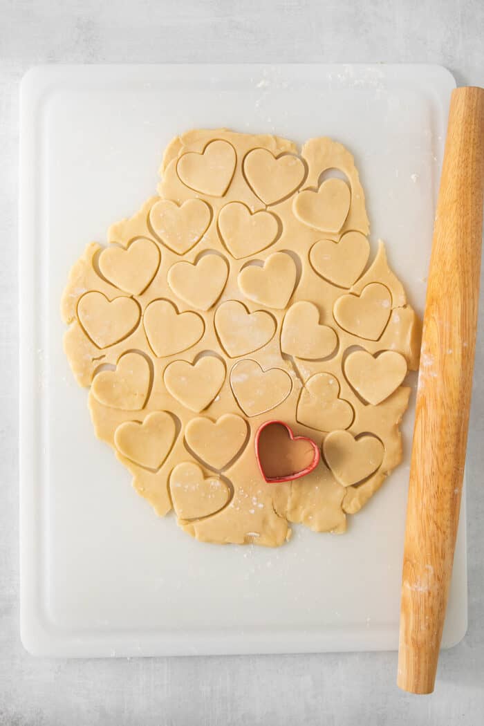 Heart shapes are cut out of a piece of cream wafer cookie dough on a white cutting board, with a rolling pin to the right.