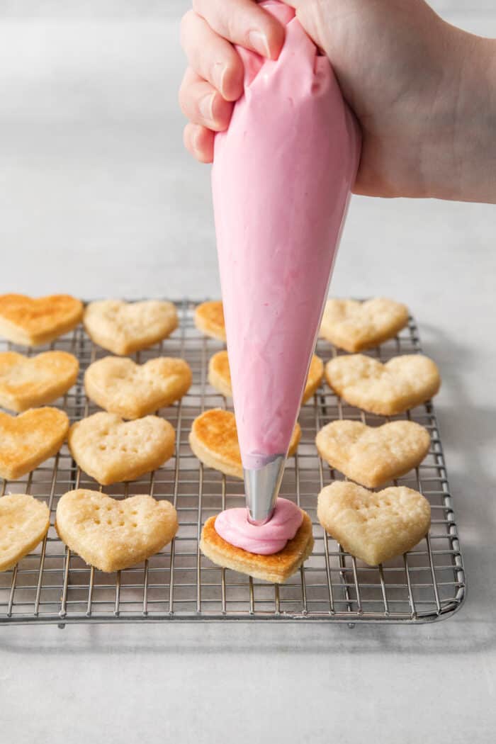 Strawberry buttercream is piped onto heart shaped cream wafer cookies on a wire rack.