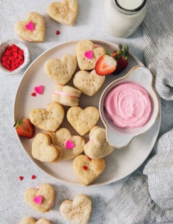 A serving dish holds strawberry cream wafer cookies decorated with heart sprinkles, with a bowl of strawberry buttercream next o them.