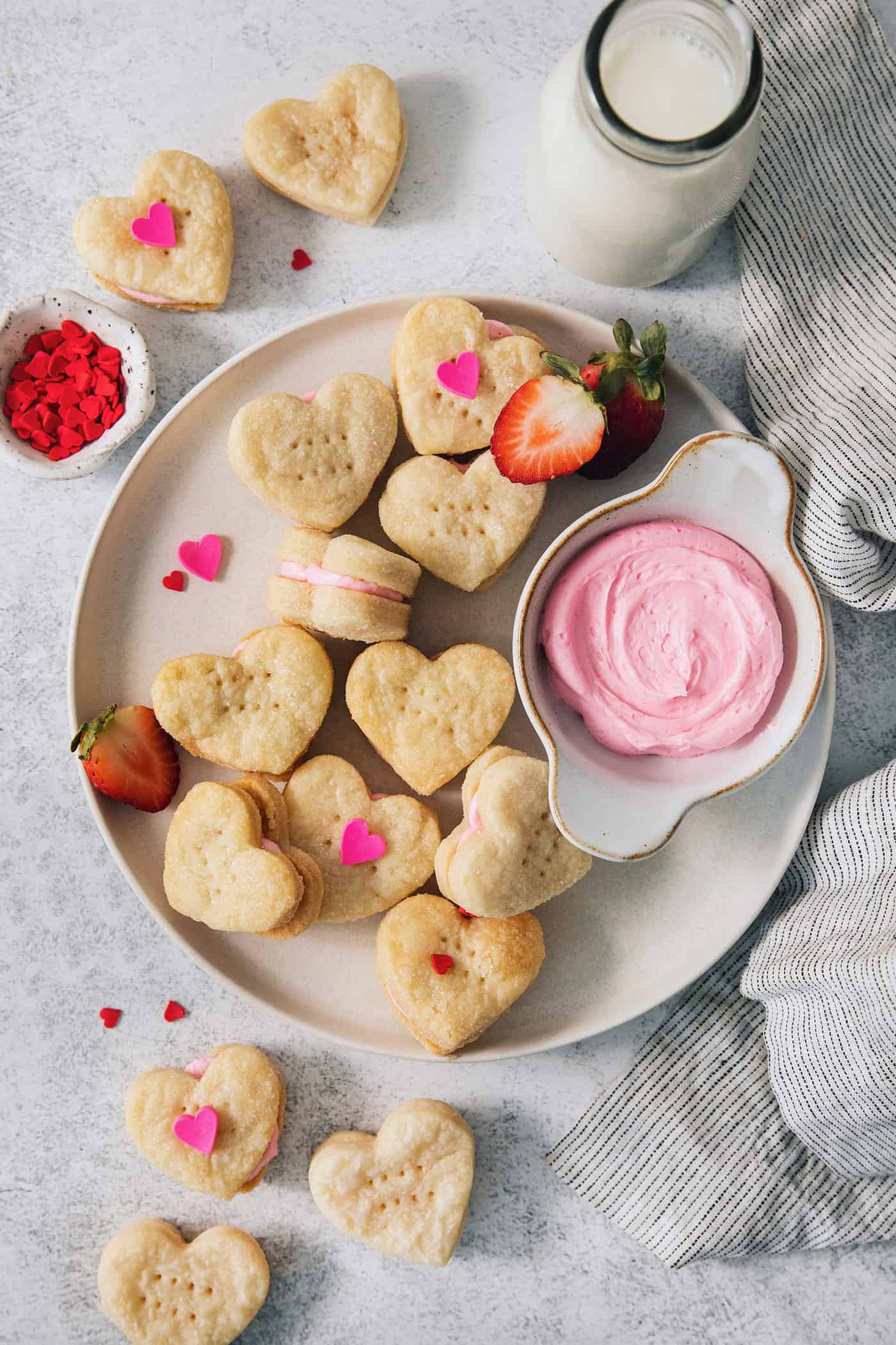 A serving dish holds strawberry cream wafer cookies decorated with heart sprinkles, with a bowl of strawberry buttercream next o them.