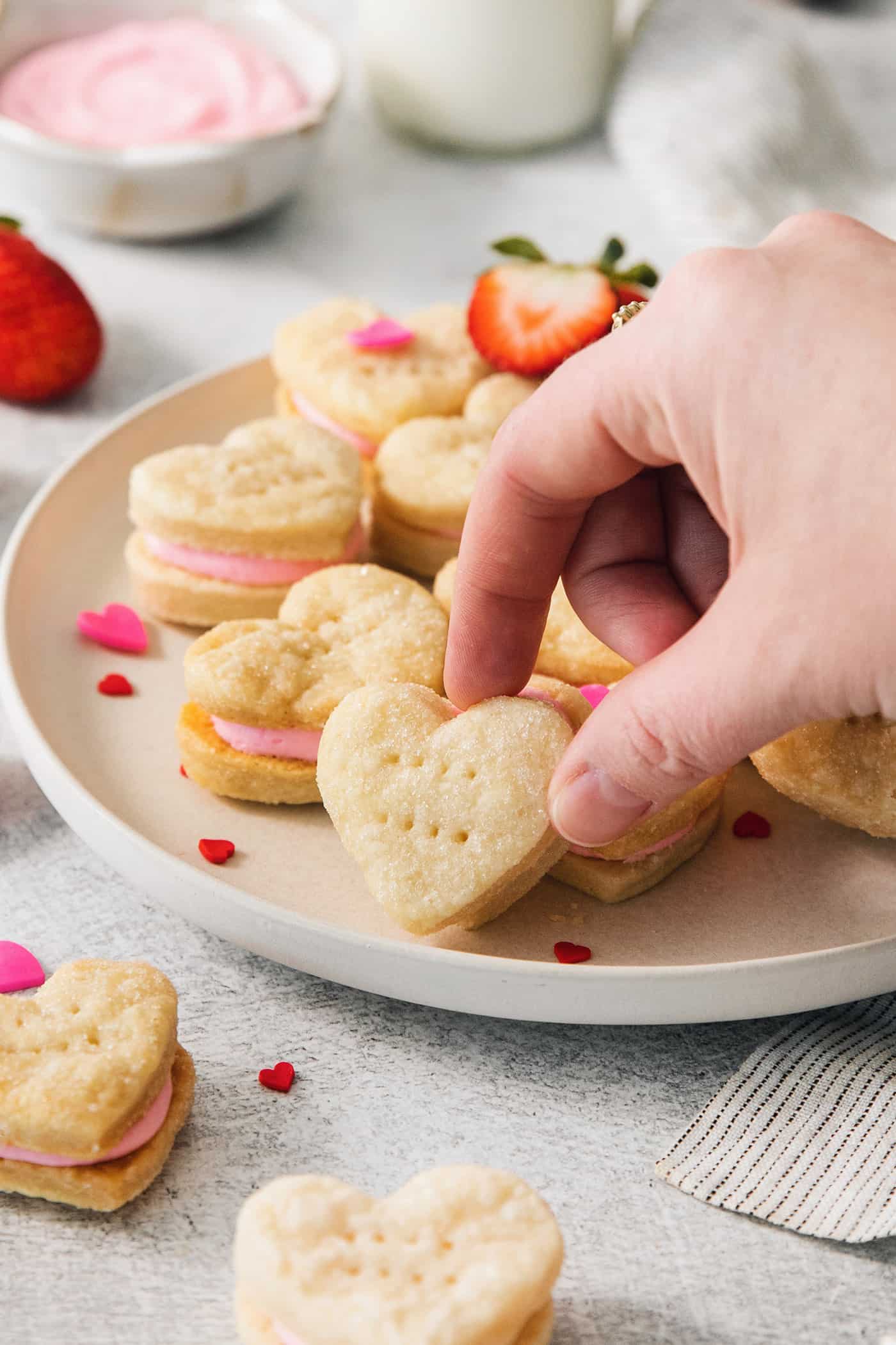 A hand reaches to pick up a heart shaped strawberry cream wafter cookie from a plate of cookies.