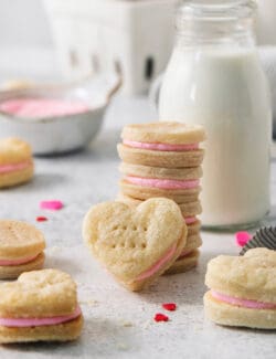 A stack of strawberry cream wafter cookies is surrounded by more cookies, with a jug of milk and a bowl of pink buttercream in the background.