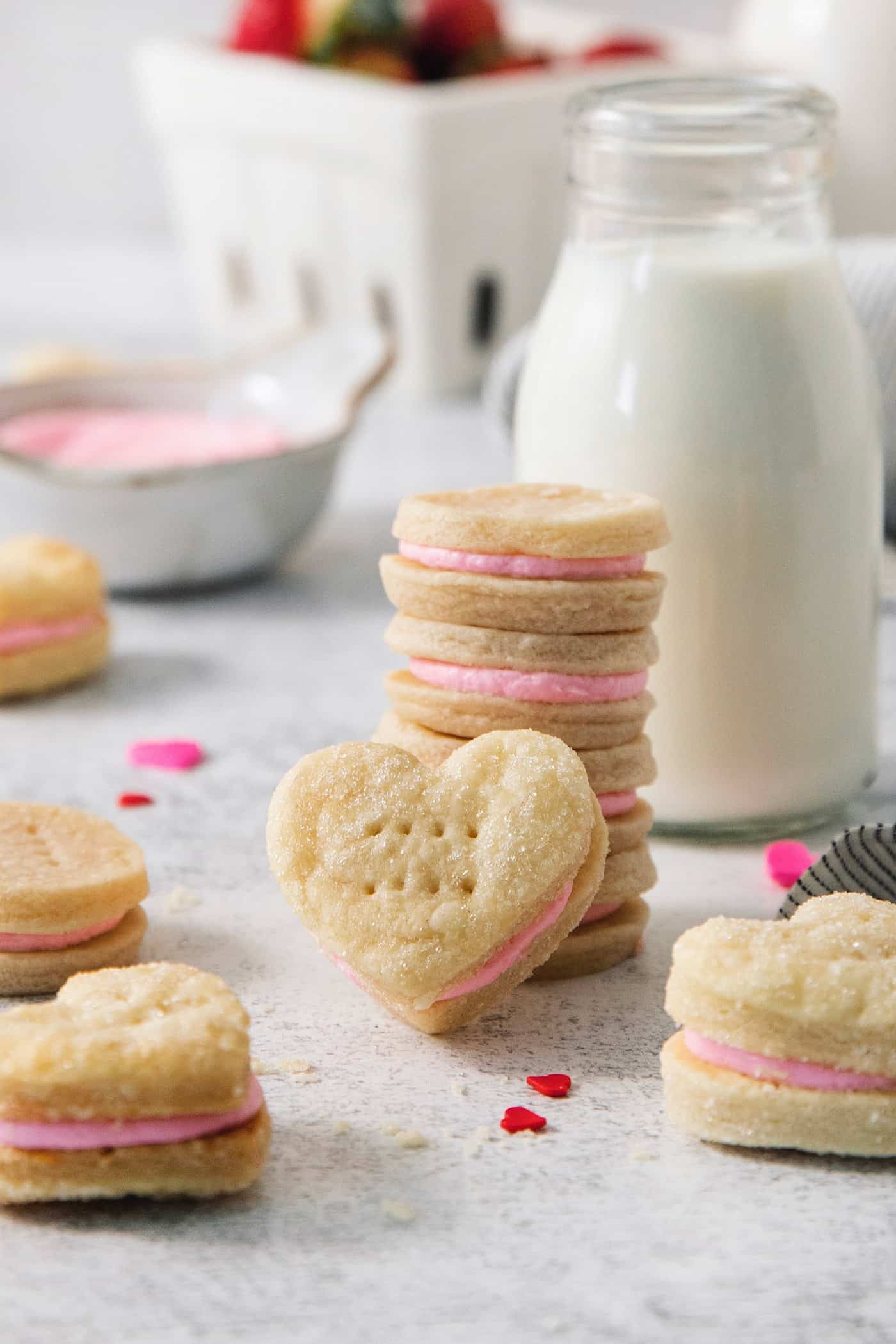 A stack of strawberry cream wafter cookies is surrounded by more cookies, with a jug of milk and a bowl of pink buttercream in the background.