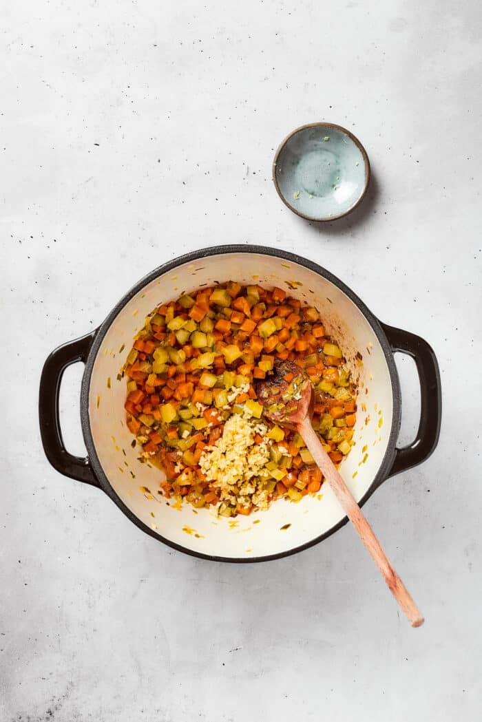 A soup base including onions, carrots, and celery cooks in a pot with a wooden spoon to stir.