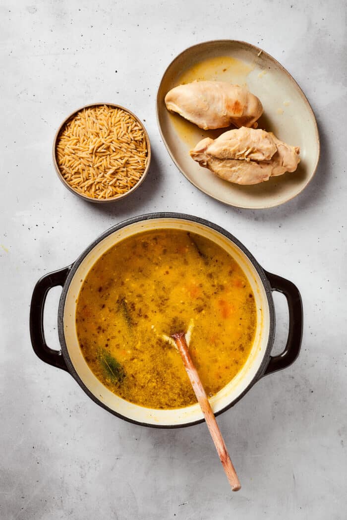 The cooked chicken is removed from the pot and placed alongside it, with a bowl of orzo waiting to be added.