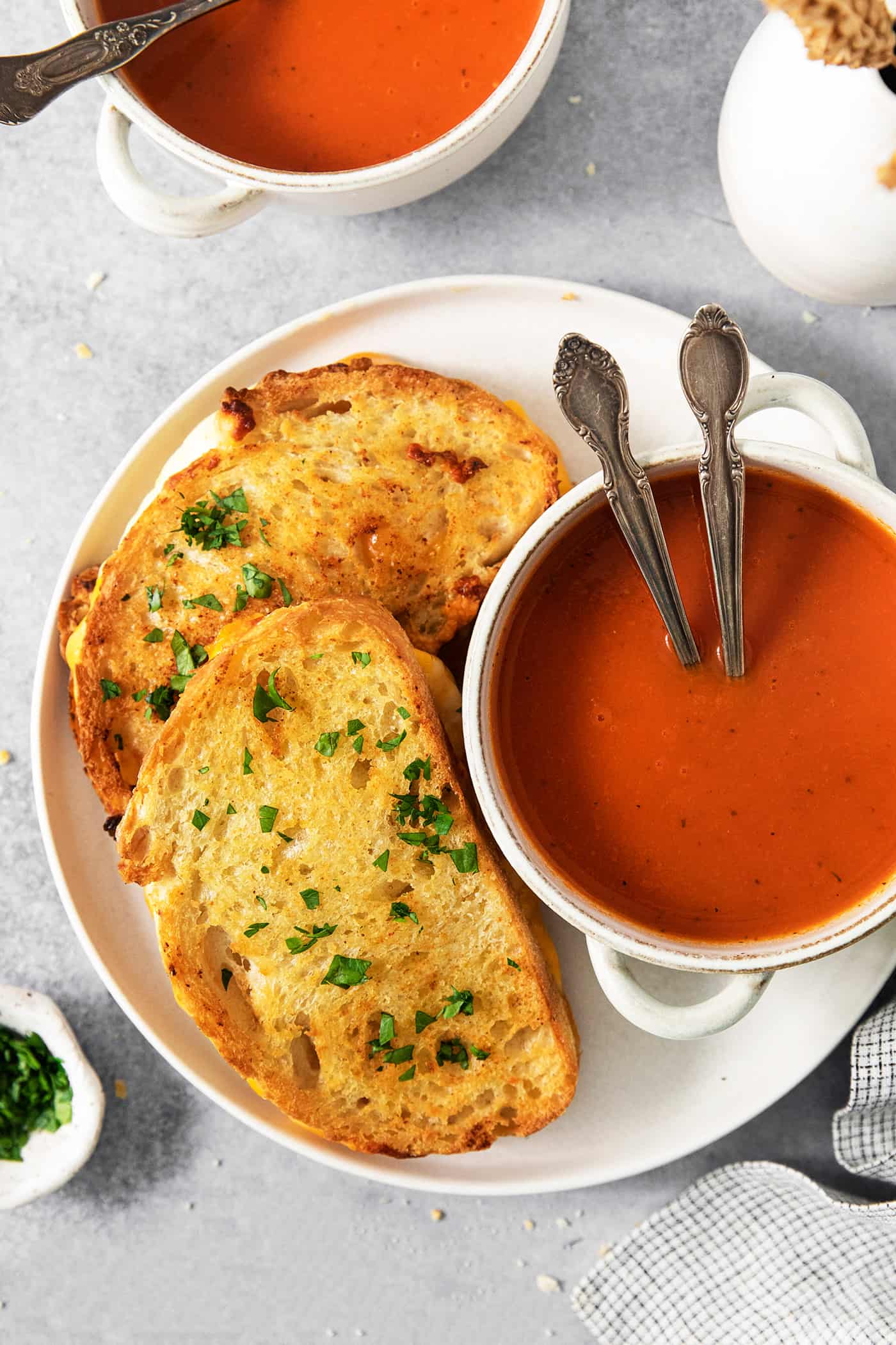 A white plate holds a small bowl of tomato soup with two spoons, and two air fryer grilled cheese sandwiches.