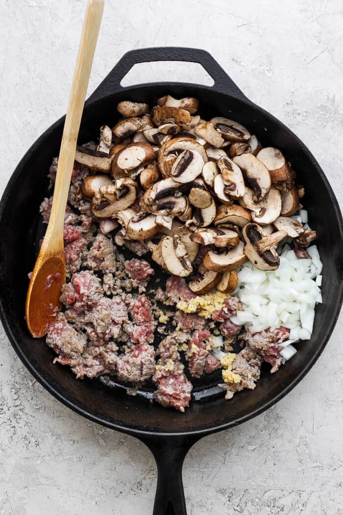 Mushrooms, sausage, onions, and garlic are stirred with a wooden spoon as they cook in a cast iron skillet.