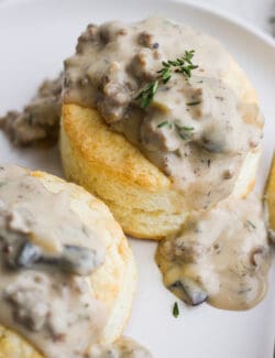 Two cream biscuits on a white plate topped with sausage and mushroom gravy.