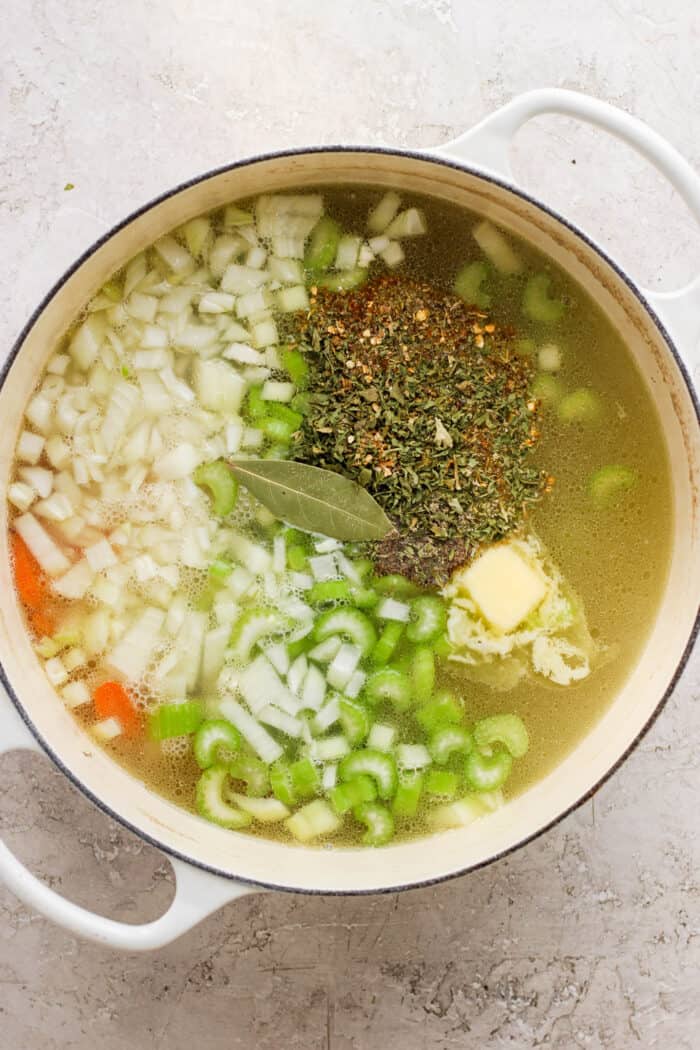 Chopped carrots, onion, celery, garlic, and herbs are shown on top of a pot of chicken broth.