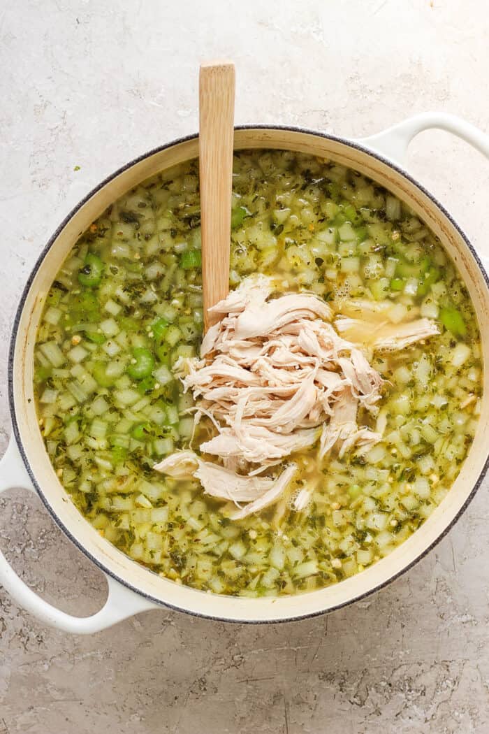 A wooden spoon stirs shredded chicken into a pot of chicken noodle soup.