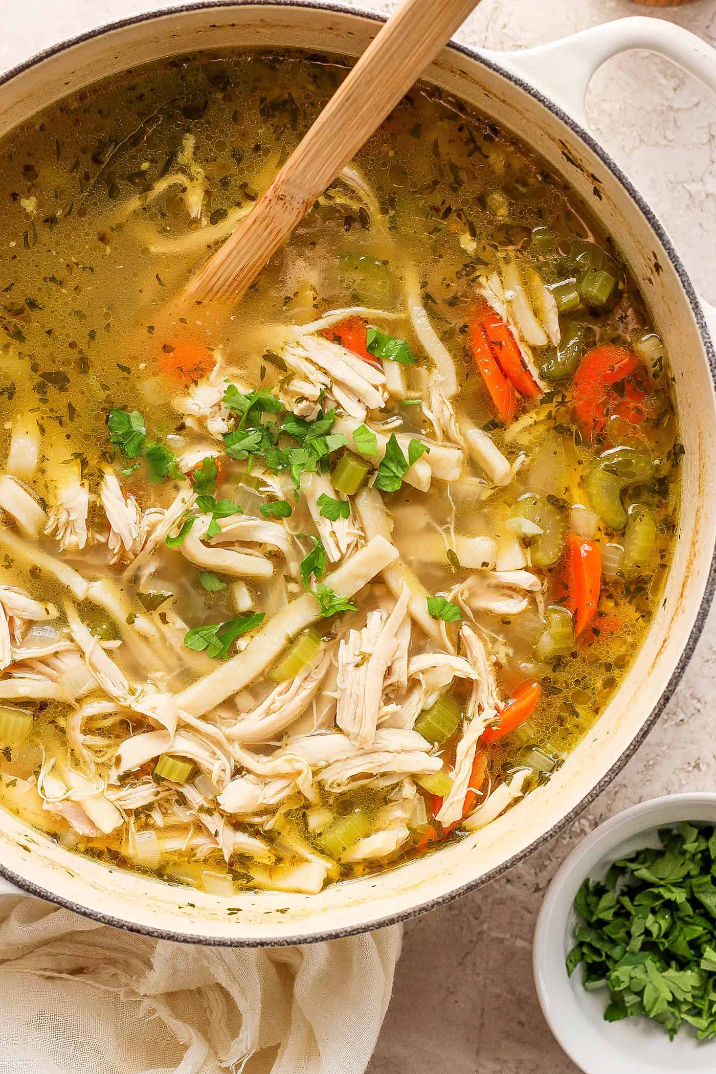 Shredded chicken, herbs and carrots are shown being stirred in a pot of chicken noodle soup.