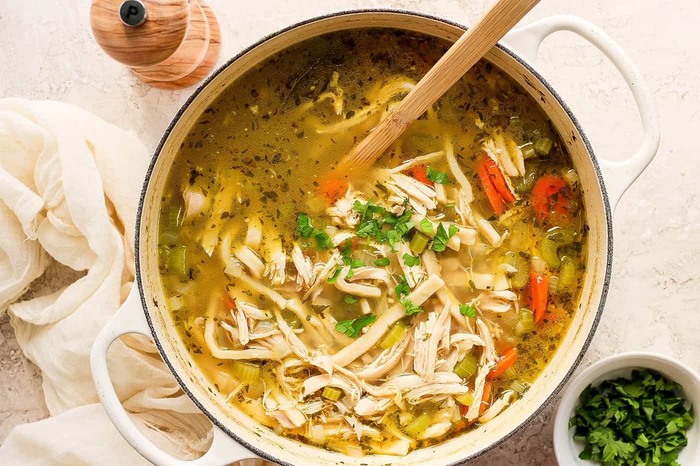 Shredded chicken, herbs and carrots are shown being stirred in a pot of chicken noodle soup.