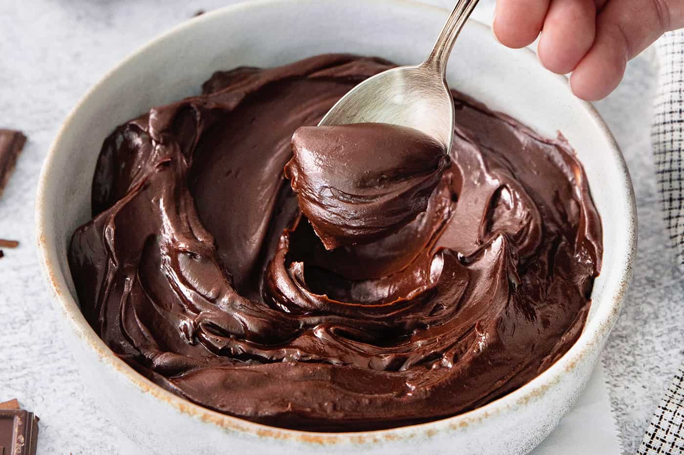 A hand holding a silver spoon stirs a white bowl of chocolate ganache.