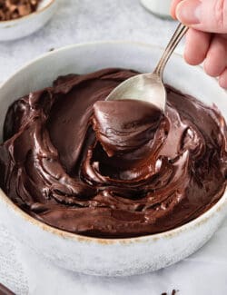 A hand holding a silver spoon stirs a white bowl of chocolate ganache.