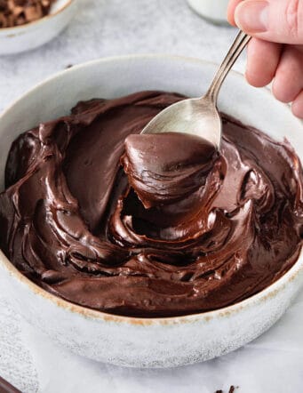 A hand holding a silver spoon stirs a white bowl of chocolate ganache.