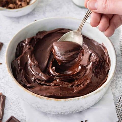 A hand holding a silver spoon stirs a white bowl of chocolate ganache.