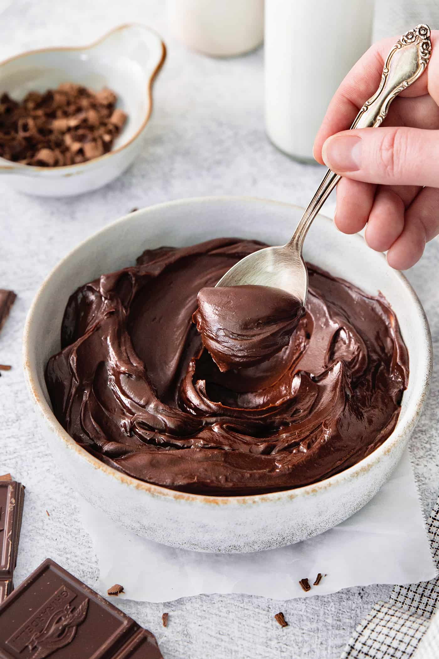 A hand holding a silver spoon stirs a white bowl of chocolate ganache.