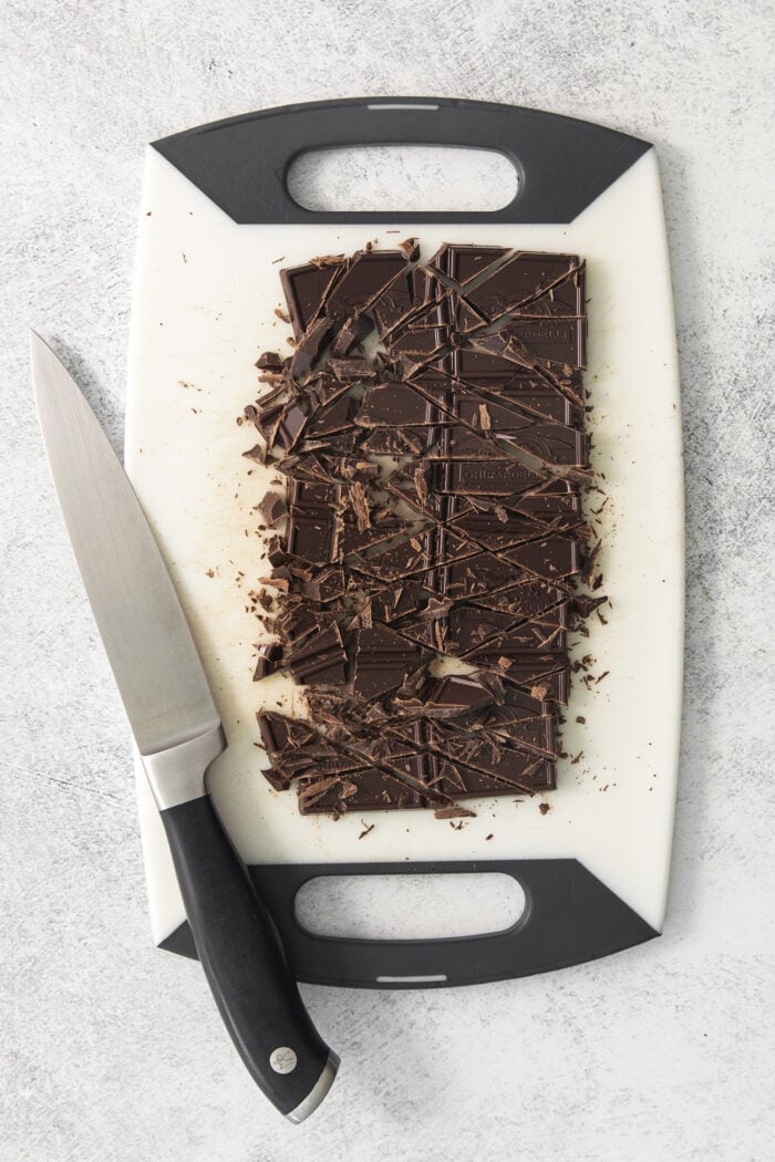 A white cutting board holds chopped chocolate with a knif resting on its left edge.