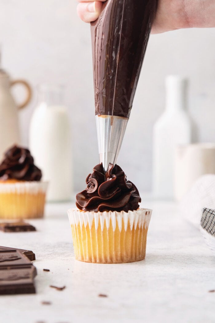A hand uses a piping bag to top a vanilla cupcake with chocolate ganache frosting.