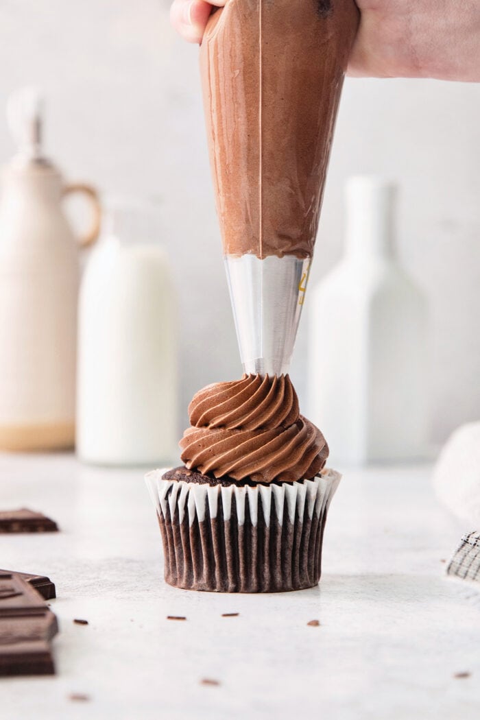 A hand pipes chocolate ganache on top of a chocolate cupcake.