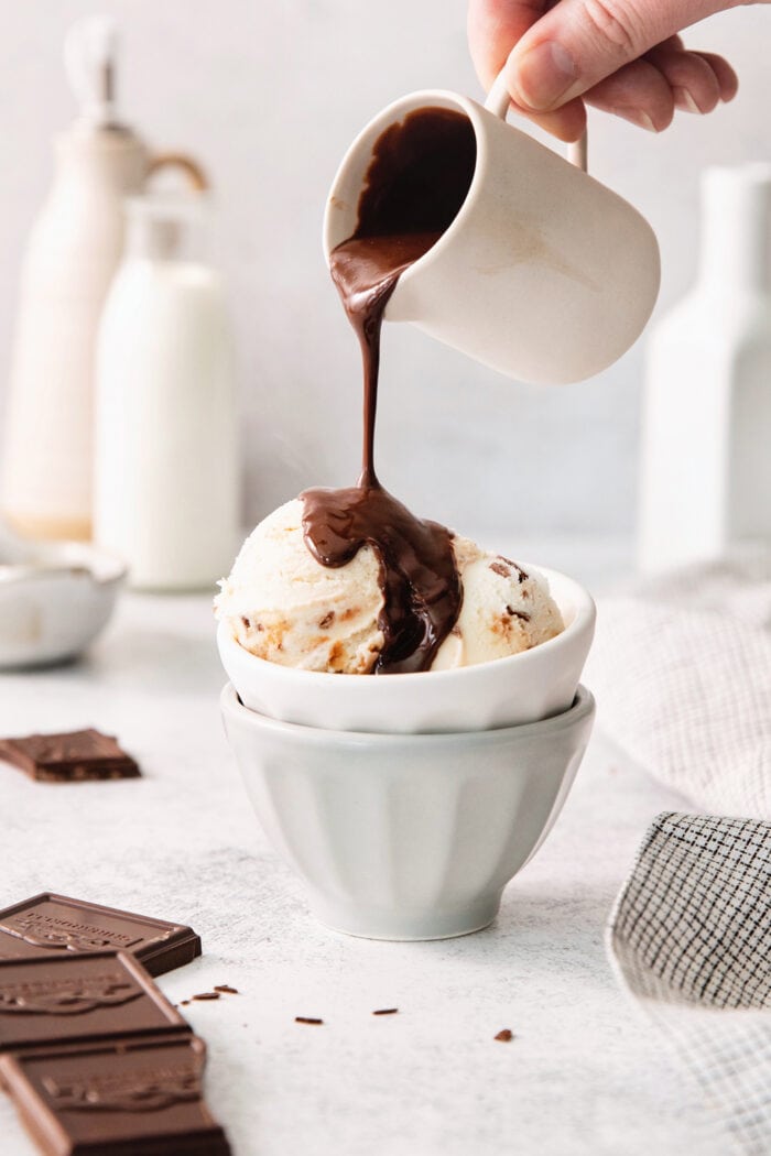 A hand holds a white pitcher and drizzles chocolate ganache over scoops of ice cream in a bowl.