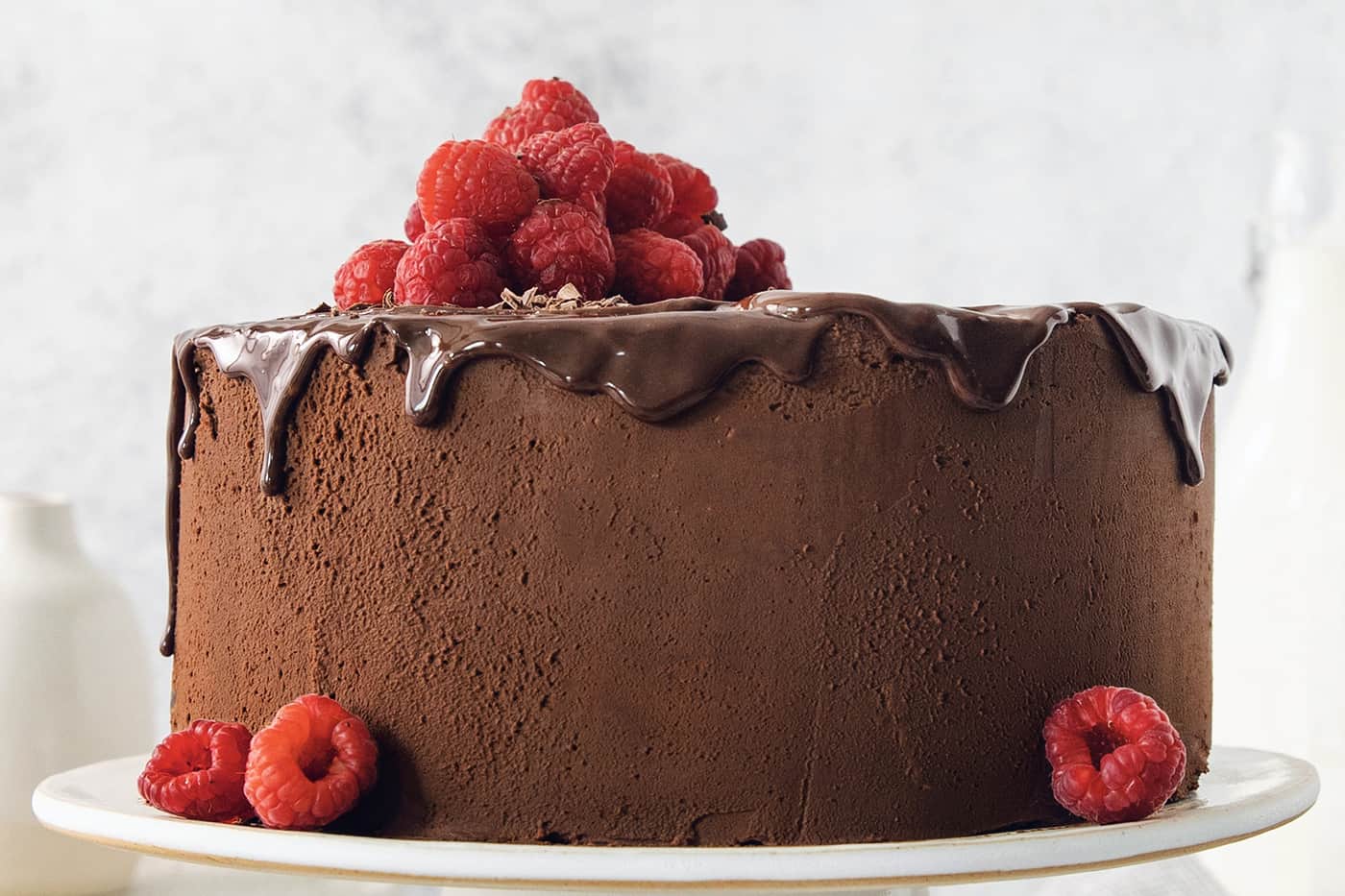 A filled and frosted two-layer chocolate ganache cake is topped with raspberries and served on a white cake stand.