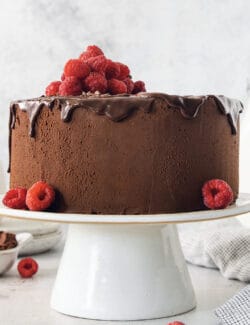 A filled and frosted two-layer chocolate ganache cake is topped with raspberries and served on a white cake stand.
