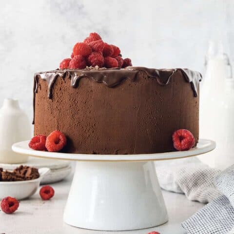 A filled and frosted two-layer chocolate ganache cake is topped with raspberries and served on a white cake stand.