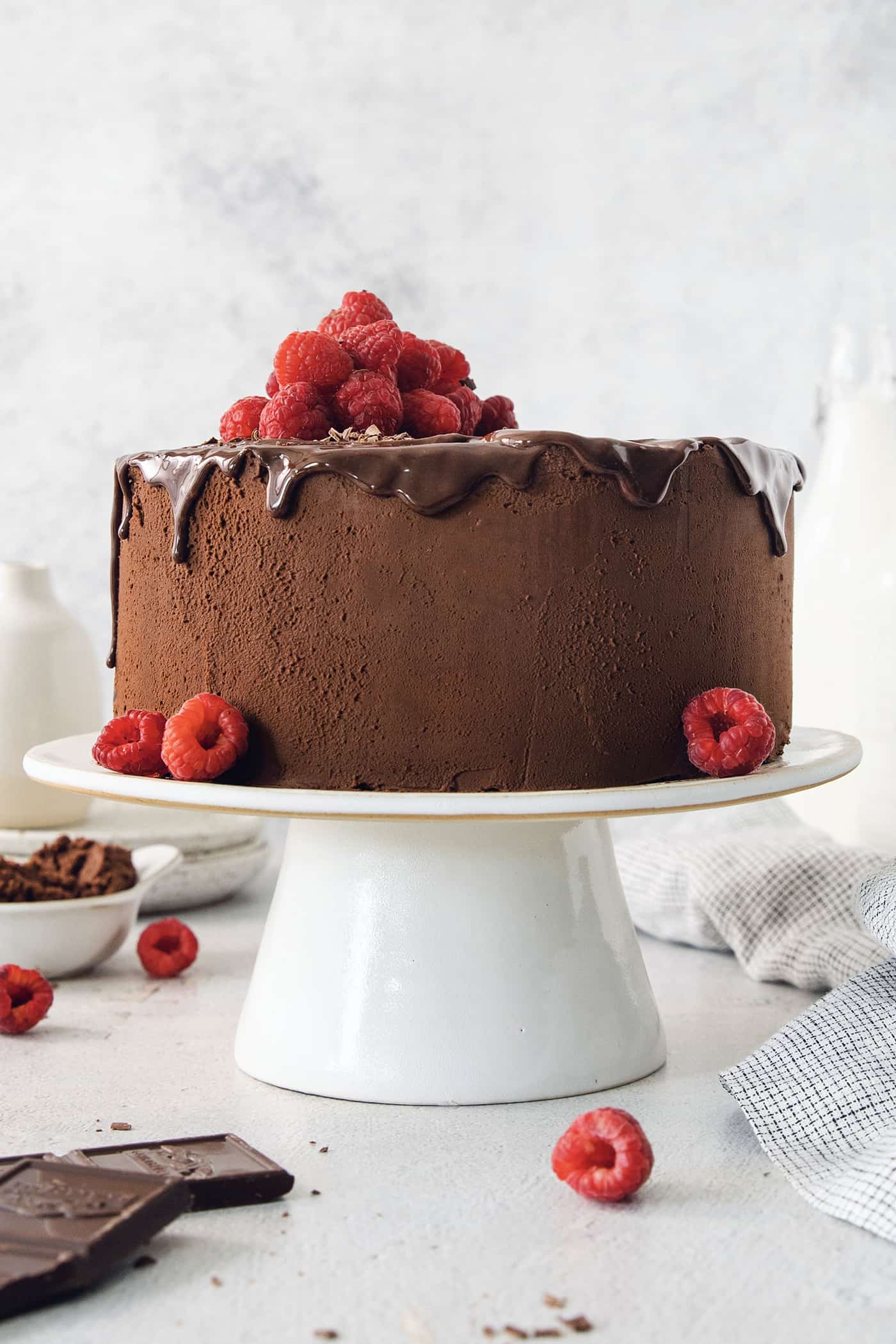 A filled and frosted two-layer chocolate ganache cake is topped with raspberries and served on a white cake stand.