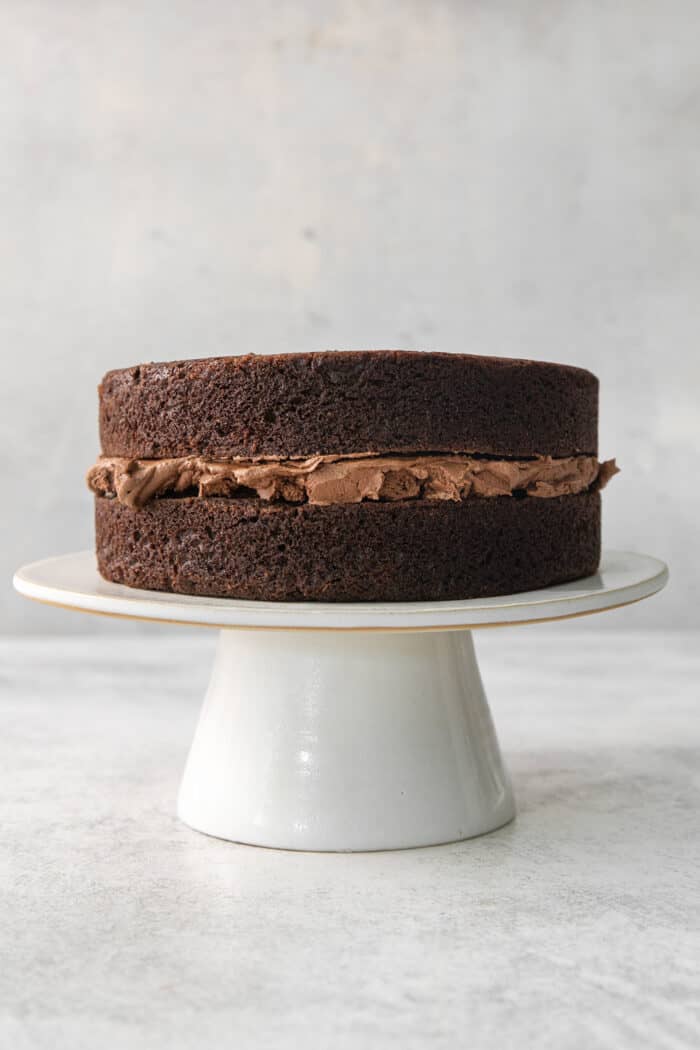 Two layers of chocolate ganache cake are filled with chocolate ganache and shown on a white cake stand.