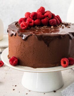 A chocolate ganache cake topped with ganache drizzle and raspberries on a white cake stand.