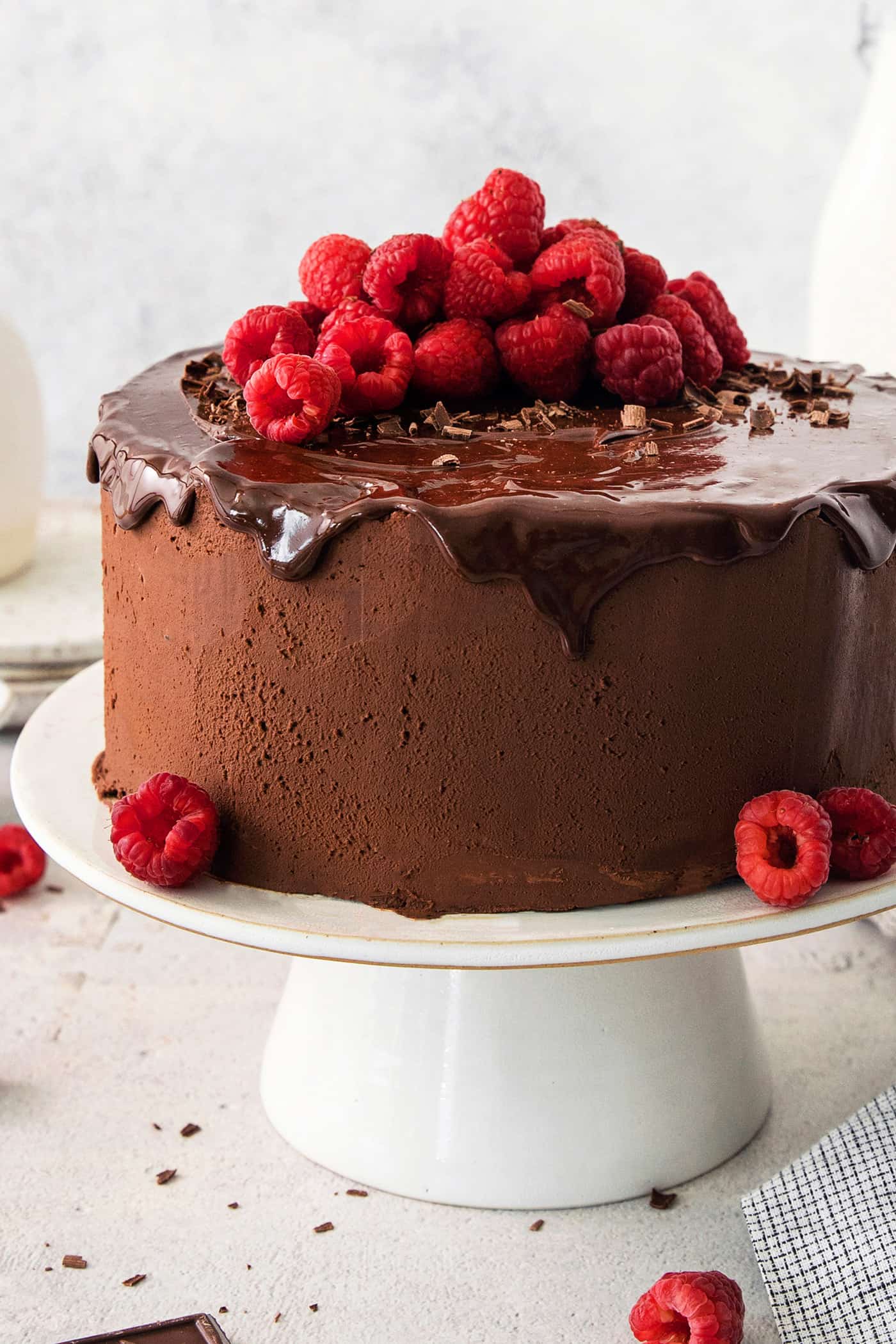 A chocolate ganache cake topped with ganache drizzle and raspberries on a white cake stand.