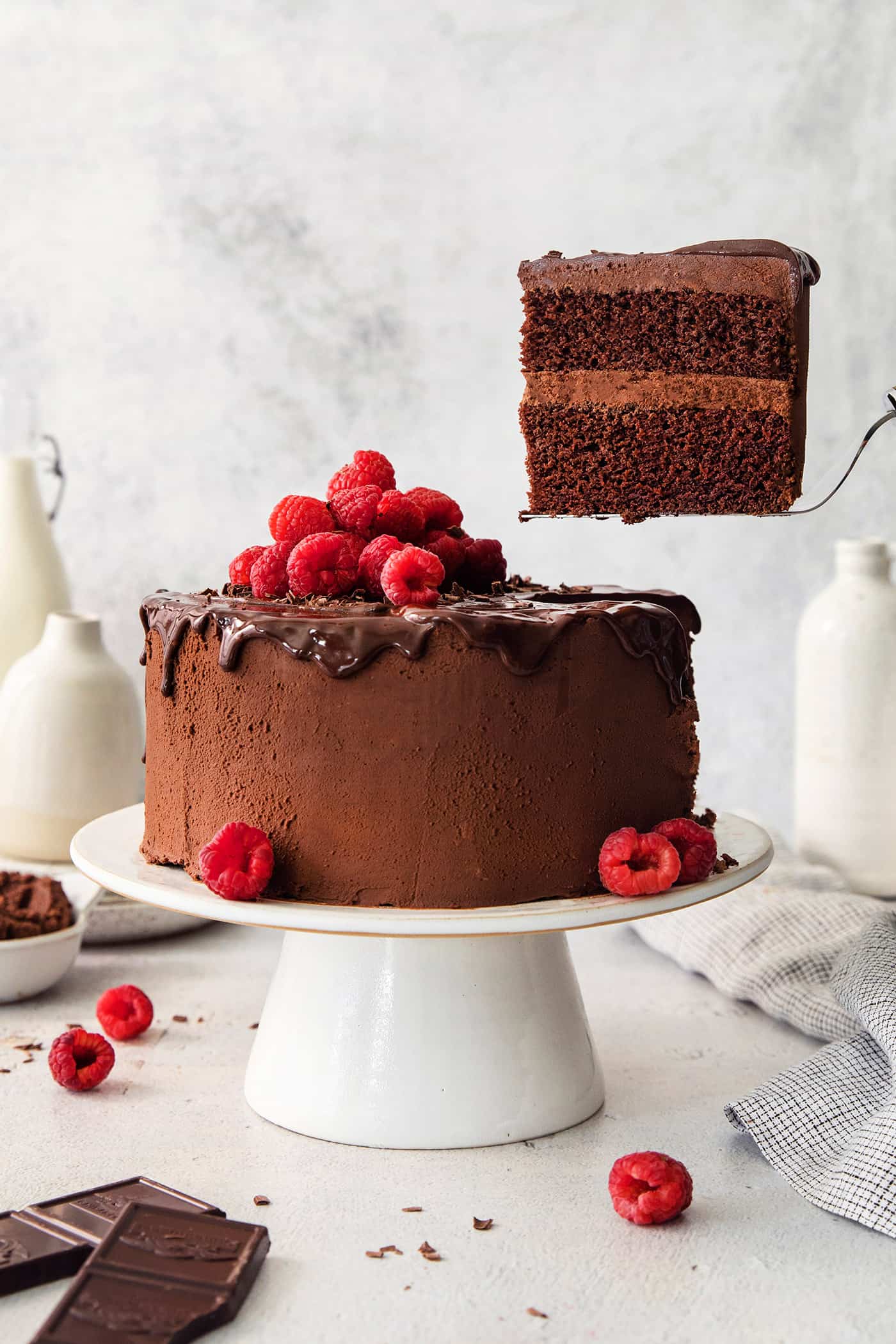 A piece of chocolate ganache cake is lifted out with a cake server from the cake on a white cake stand.