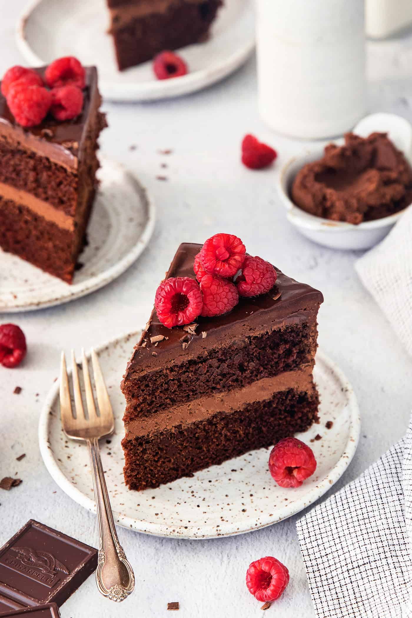 Raspberry-topped slices of chocolate ganache cake are shown on white plates on a table.