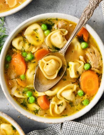 A bowl of lemon chicken tortellini soup on a table surrounded by more bowls of soup, peas, and lemons.