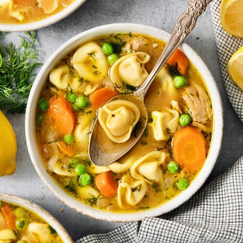 A bowl of lemon chicken tortellini soup on a table surrounded by more bowls of soup, peas, and lemons.
