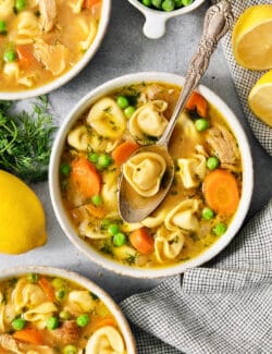 A bowl of lemon chicken tortellini soup on a table surrounded by more bowls of soup, peas, and lemons.