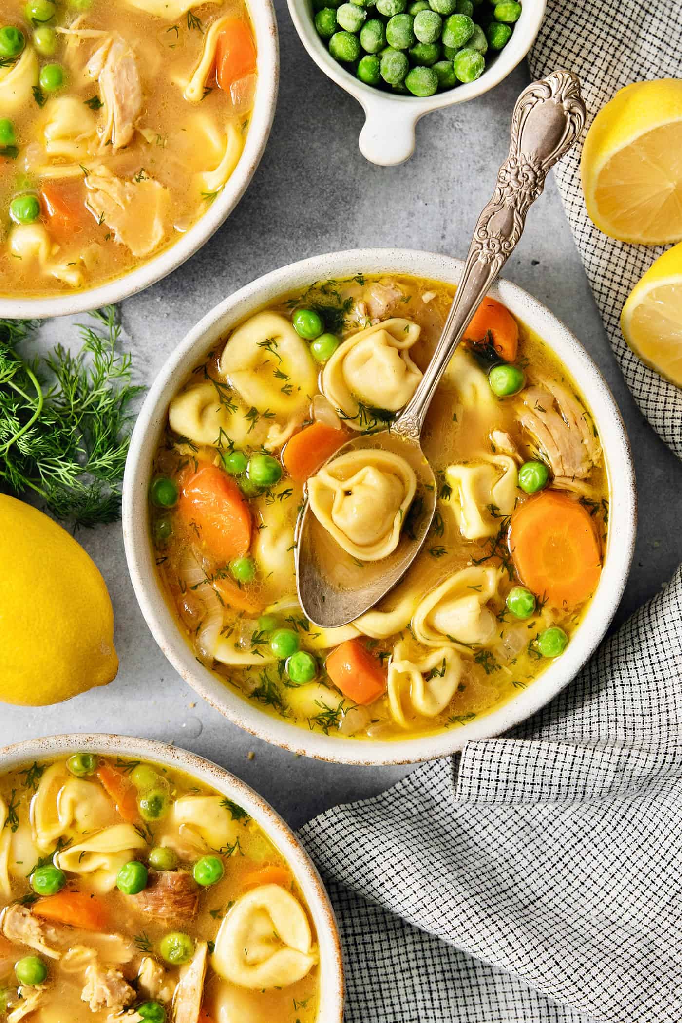 A bowl of lemon chicken tortellini soup on a table surrounded by more bowls of soup, peas, and lemons.