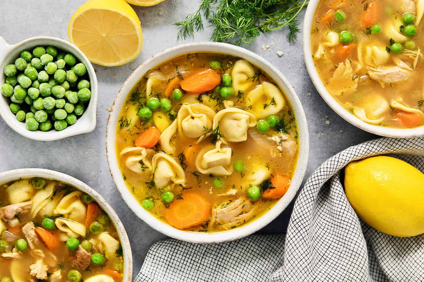 A bowl of lemon chicken tortellini soup on a table surrounded by more bowls of soup, peas, and lemons.