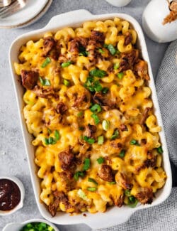 A cheesy casserole dish full of pulled pork mac and cheese on a table with a dish of chopped green onions nearby.