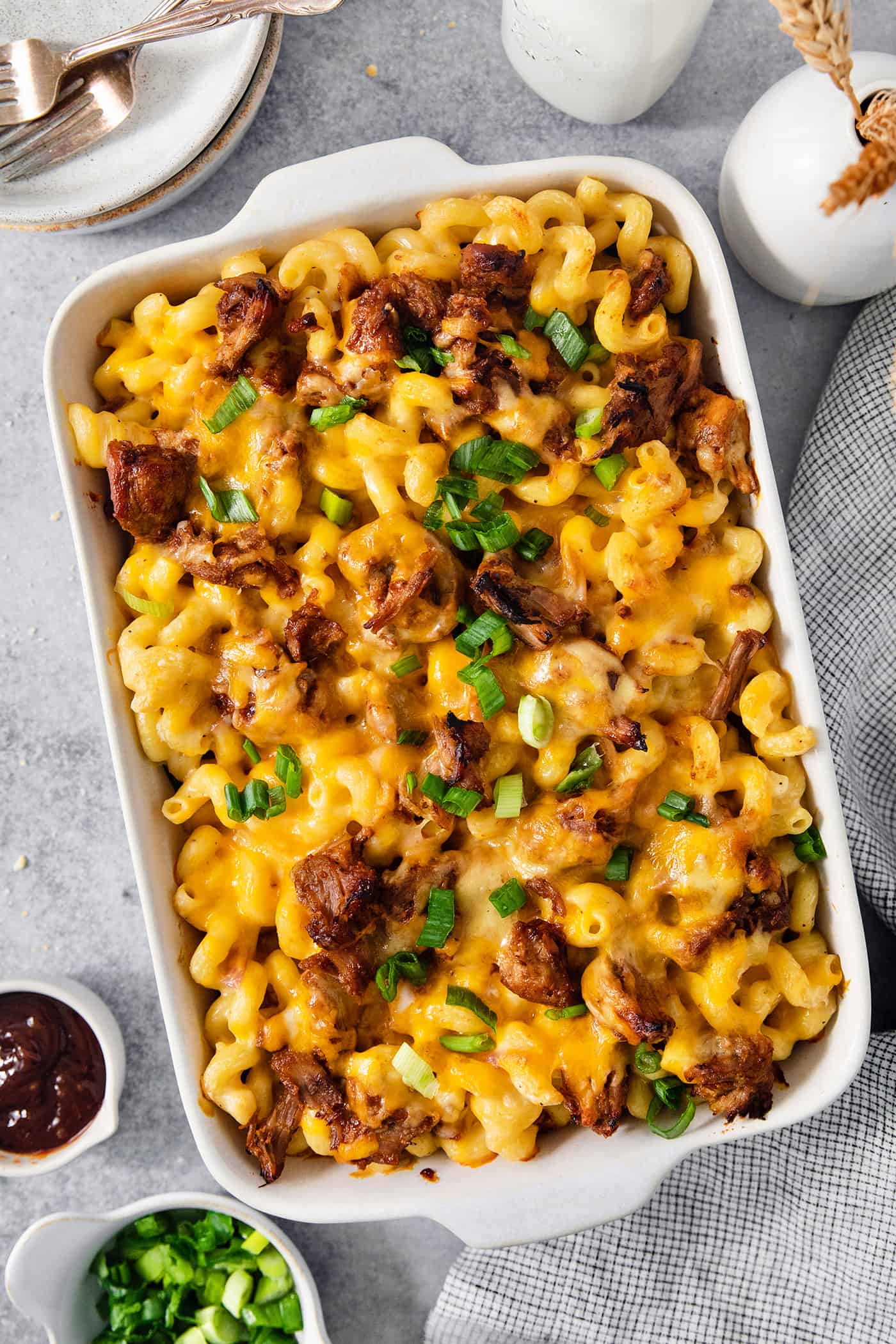 A cheesy casserole dish full of pulled pork mac and cheese on a table with a dish of chopped green onions nearby.